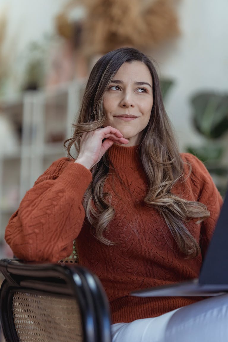 Thoughtful positive female freelancer wearing casual clothes sitting on armchair and using netbook and looking away